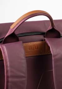 Zaino bordeaux con una superficie testurizzata, manico in pelle e logo inciso su un'etichetta in pelle marrone. Progettato con spallacci regolabili.