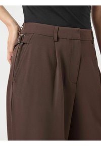 Primo piano di pantaloni marroni a vita alta con fibbia laterale, pieghe e tasche, abbinati a un top nero, che mostrano l'area della vita e dei fianchi.