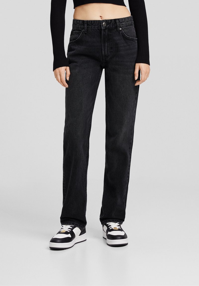 Personne portant un jean noir taille haute à jambes droites, des baskets noires et blanches, et un haut noir court à manches longues, debout devant un fond gris.