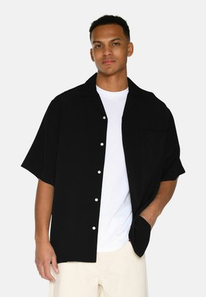 Black short-sleeve shirt with a loose fit, button front, pocket on the left chest, worn over a white t-shirt. Soft, smooth texture.
