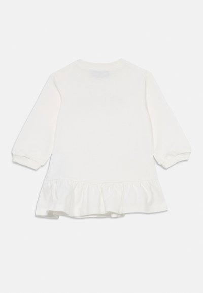 Vit långärmad babydress med volang i nederkant och elastiska manschetter, visad bakifrån mot en enkel bakgrund.