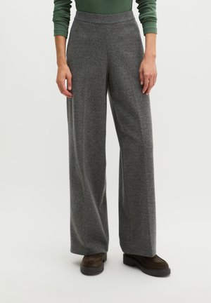 Persoon draagt grijze wijde broek, groene top met lange mouwen en donkerbruine schoenen, staand tegen een effen lichte achtergrond.