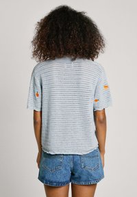 Top corto a maniche corte di colore blu chiaro in maglia con un motivo testurizzato e dettagli ricamati arancioni sulle maniche, abbinato a pantaloni corti di jeans.