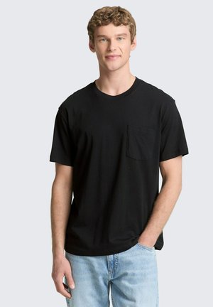 Młody mężczyzna z kręconymi włosami, ubrany w czarny t-shirt z kieszeniami i jasnoniebieskie dżinsy, stojący z jedną ręką w kieszeni na jednolitym tle.