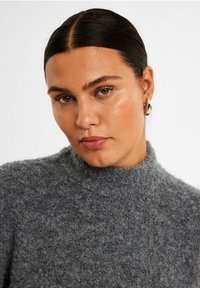 Grå strikket sweater med høj hals, struktureret stof og et blødt, fl fluffy udseende. Modellen bærer subtile øreringe og naturlig makeup.