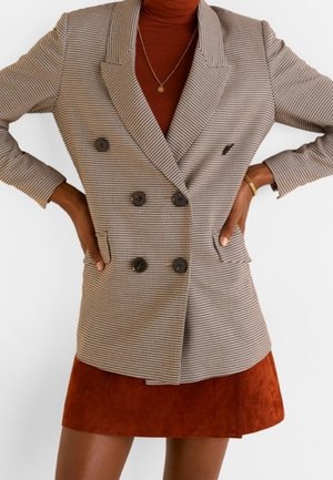 Frau trägt ein beige kariertes, doppelt geknöpftes Blazer über einem rostfarbenen Rollkragenpullover und einem rostfarbenen Wildleder-Minirock, die Hände in die Hüften gestützt, mit Goldarmband und Halskette.