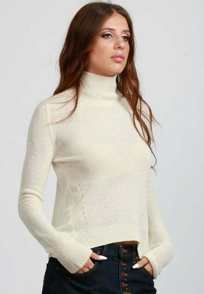 Donna con lunghi capelli castani, che indossa un maglione a collo alto color crema e jeans blu scuro, con le mani nelle tasche, che guarda leggermente verso destra.