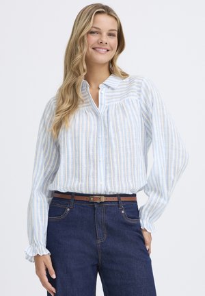 Lächelnde Frau mit welligem, blonden Haar, die eine blau-weiß geringelte Bluse mit langen Ärmeln trägt, die in eine dunkelblaue Jeans mit einem braunen Gürtel gesteckt ist.