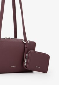 Borsa a tracolla bordeaux con manici doppi, dotata di accessori in ottone argentato e un piccolo portafoglio attaccato. Design minimalista con logo impresso.