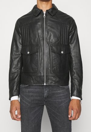 Schwarze Lederjacke mit Frontreißverschluss, Klappentaschen mit silbernen Knöpfen, getragen von einer Person in dunkelgrauen Jeans vor einfarbigem Hintergrund.