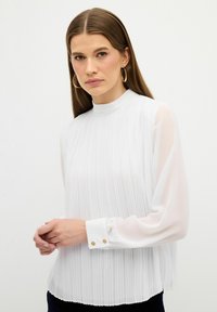 Weiße, gefaltete Bluse mit hohem Kragen, langen transparenten Ärmeln und geknöpften Manschetten mit goldenen Akzenten. Weicher, leichter Stoff.