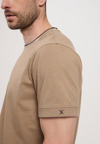 Beżowy T-shirt z okrągłym dekoltem i krótkimi rękawami, wykonany z miękkiej bawełny. Posiada granatowy akcent na kołnierzyku oraz ciemny haft w kształcie X na rękawie.