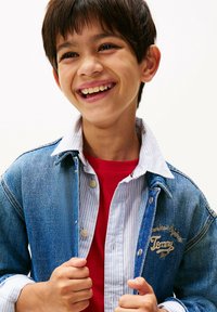 Chaqueta de mezclilla con un lavado azul, camisa a rayas azul claro debajo, camiseta roja visible, con logo bordado en la chaqueta.
