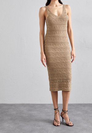 Kvinde iført en ærmeløs, knælængde beige hæklekjole med V-hals og sorte høje hæle med stropper, stående på en grå gulv.