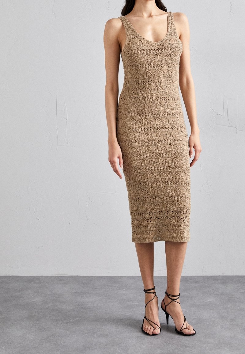 Femme portant une robe beige en crochet sans manches, longueur genoux, avec un col en V et des sandales noires à talons hauts avec des brides, debout sur un sol gris.