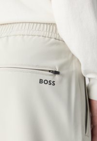 Pantalon de jogging gris clair avec une taille élastique, une poche zippée et un logo "BOSS" noir sur le côté. Texture de tissu lisse et douce.