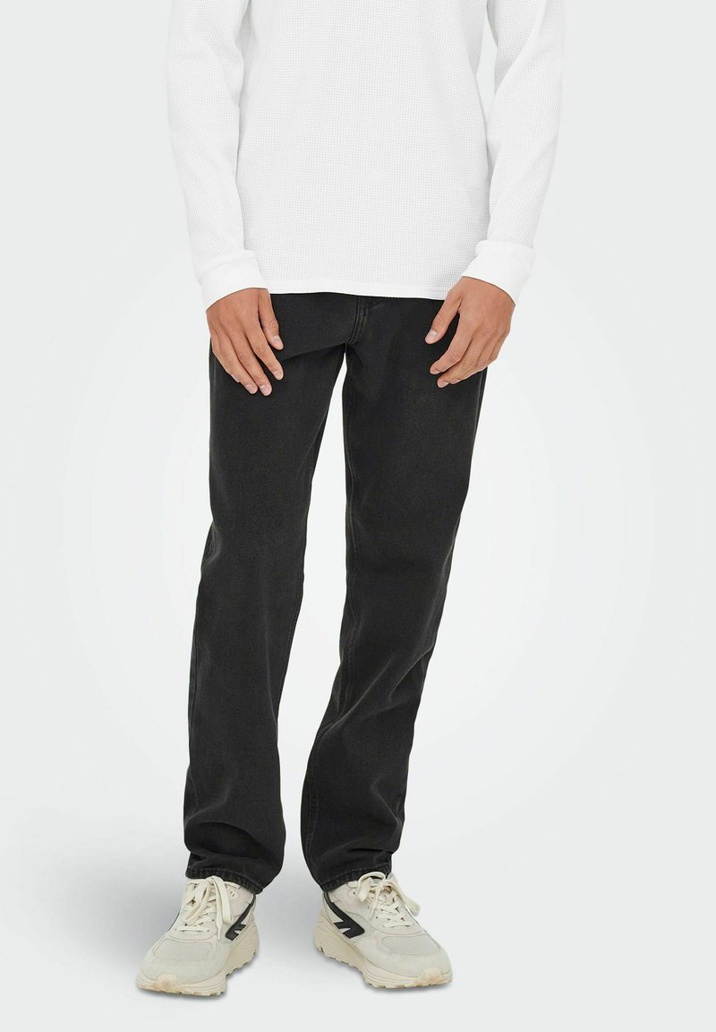Jeans negros de corte recto, con una textura suave. Combinados con una camisa blanca de manga larga y zapatillas de color claro.