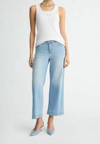 Mujer vistiendo una blusa sin mangas blanca, vaqueros de pierna ancha azul claro y tacones de aguja azul sobre un fondo liso.