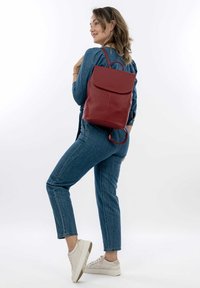 Un sac à dos en cuir rouge avec une fermeture à rabat, une surface texturée, des bretelles réglables et une poignée supérieure, porté par une personne en denim.