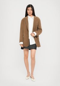 Donna in piedi con indosso un blazer oversize marrone, camicia bianca con bottoni, gonna scura e scarpe mules bianche a punta, su uno sfondo neutro.