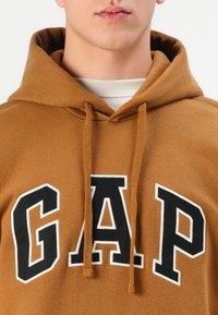 Personne portant un sweat à capuche marron avec des lettres "GAP" en noir et blanc sur le devant et des cordons qui pendent.
