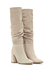 Stivali in suede beige al ginocchio con design slouchy, punta a punta e tacco a blocco. Presentano una cerniera laterale per la chiusura.