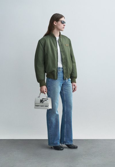 Γυναίκα που φοράει πράσινο μπλούζα τύπου bomber, λευκό τοπ, μπλε τζιν, μαύρα τακούνια, γυαλιά ηλίου, κρατώντας λευκή τσάντα χειρός με την επιγραφή «Εισαγάγετε Χρήματα Εδώ».