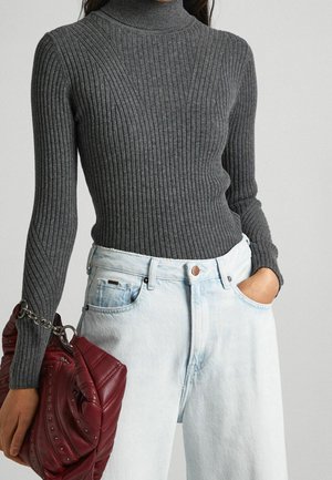 Femme portant un pull à col roulé côtelé gris, un jean bleu clair, tenant un sac à main matelassé rouge plié avec des clous en métal dans une main.
