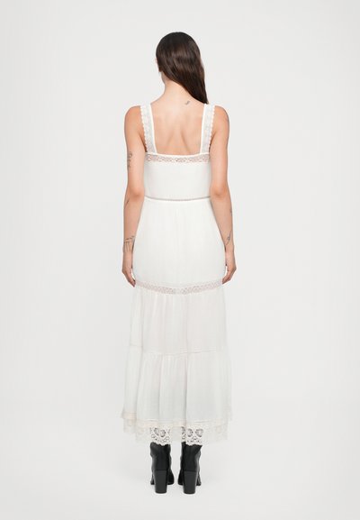 Vestido maxi blanco con tirantes de encaje, tejido texturizado y diseño en capas. Presenta un borde de encaje en el dobladillo. Se lleva con botas de tacón negras.