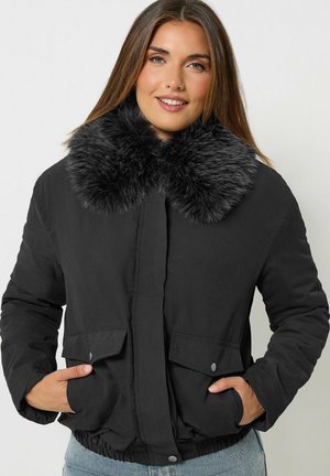 Jeune femme portant une veste d'hiver noire avec un grand col en fourrure noire, mains dans les poches, souriante, sur un fond clair uni.