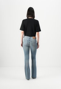 Woman standing facing away wearing black short-sleeve shirt and light blue flared jeans against white background.