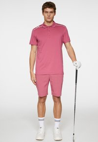 Rosa polo-tröja med vita axelränder, matchad med kortbyxor. Vita sportstrumpor med svarta ränder och vita sneakers. Golfklubba i handen.