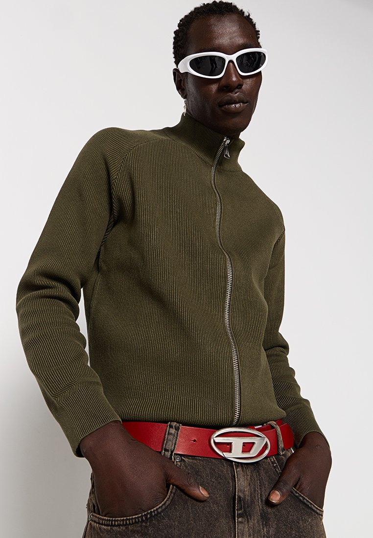 Maglione a zip in cotone ribbed verde oliva, cintura rossa con fibbia argentata, pantaloni in denim neri e occhiali da sole bianchi. Design semplice e aderente.