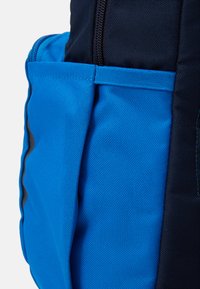 Blue fabric backpack with a textured finish, featuring a side pocket in a lighter shade of blue and dark navy accents. Zipper closure visible.