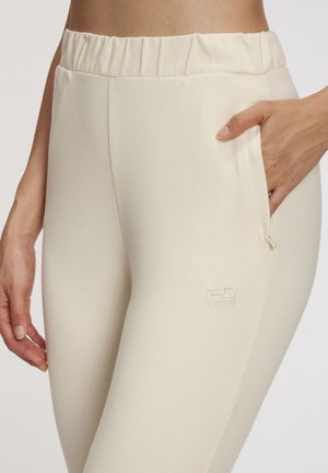 Beige elastische taillebroek met een gladde textuur, voorzien van zijzakken en een subtiele geborduurde logo op de bovenbeen.
