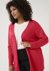 Cardigan rouge avec des manches longues, ouverture à l'avant et tissu doux. Porté sur un haut blanc et associé à un pantalon foncé.