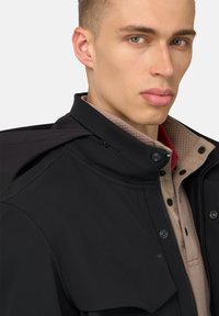 Giacca nera con zip e colletto alto, caratterizzata da una superficie liscia e bottoni a pressione, indossata sopra una maglietta in maglia beige chiaro con colletto.