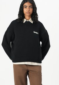 Zwarte gebreide trui met een geribde onderkant en manchetten, voorzien van een klein wit Carhartt-logo op de voorkant, gelaagd over een crèmekleurige overhemd met kraag.