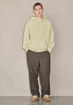 Junger Mann steht mit den Händen in den Taschen, trägt einen blassgelben Hoodie, lockere braun karierte Hose und beige Sneakers vor einfachem Hintergrund.