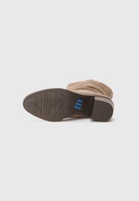 Botín de ante beige con un diseño arrugado y puntera afilada, que presenta una suela de goma marrón con un logotipo azul y un patrón decorativo.