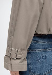 Κοντινό πλάνο μανικιού μπεζ σακακιού με λεπτομέρεια από ασημένια αγκράφα και τσέπη από μπλε τζιν με κεντημένο λογότυπο.