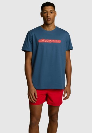 Niebieska bawełniana koszulka z krótkimi rękawami i okrągłym dekoltem, z neonowo-czerwonym logo "diverse" z przodu. Noszona z czerwonymi szortami.