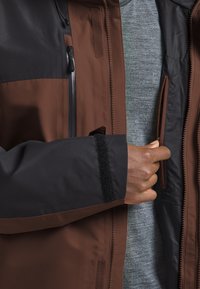 Wasserfeste Jacke in Braun und Schwarz mit einer Reißverschlusstasche, Klettverschlussakzenten und einer weichen grauen Innenlage. Sie bietet eine glatte Textur und strapazierfähiges Material.