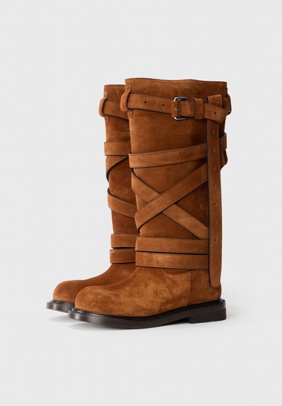 Brown suede knee-high boots with crisscross straps, buckle detail, and a smooth black sole. Matte texture with a rounded toe design.
