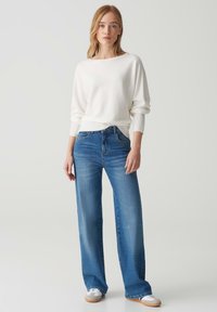 Weißer Strickpullover mit lockerem Schnitt, kombiniert mit hellblauen weiten Jeans. Weiße Sneakers mit grauen Akzenten runden das Outfit ab.