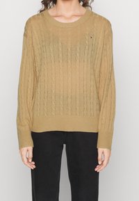 Beiger Pullover mit Zopfmuster, Rundhalsausschnitt, gerippten Bündchen und Saum; mit kleinem Logo auf der Brust. Getragen zu schwarzen Hosen.