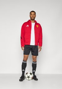 Crvena jakna na patentni zatvarač s logotipom Manchester Uniteda, bijela majica, crne kratke hlače, crne čarape s bijelim prugama i crne nogometne kopačke.