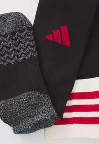 Črne in sive teksturirane nogavice z zigzag vzorci, s rdečim logotipom in belimi črtami na robu. Pletena tkanina z rebrastimi podrobnostmi.