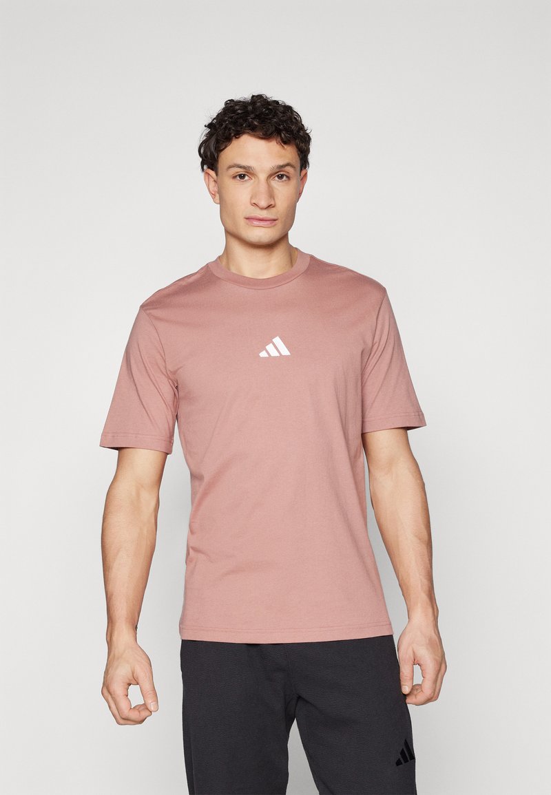 Korte mouwen, lichtroze katoenen t-shirt met een ronde hals en witte logodetail op de borst; gecombineerd met donkere broeken.