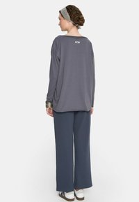 Kobieta z warkoczami i szarym opaską na głowie, nosząca luźną ciemnoszary bluzkę, dopasowane szerokie spodnie, bransoletki i białe sneakersy, odwrócona tyłem.